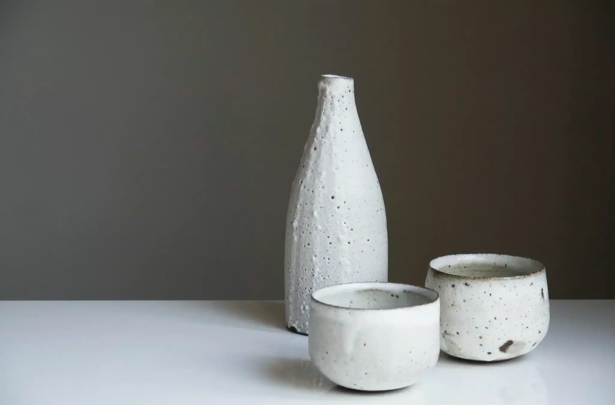 A minimalist display of ceramic art consisting of a tall, textured white vase alongside two matching bowls on a white surface. The items, featuring a speckled pattern reminiscent of natural stone, are set against a gray background that accentuates their simple yet elegant forms.