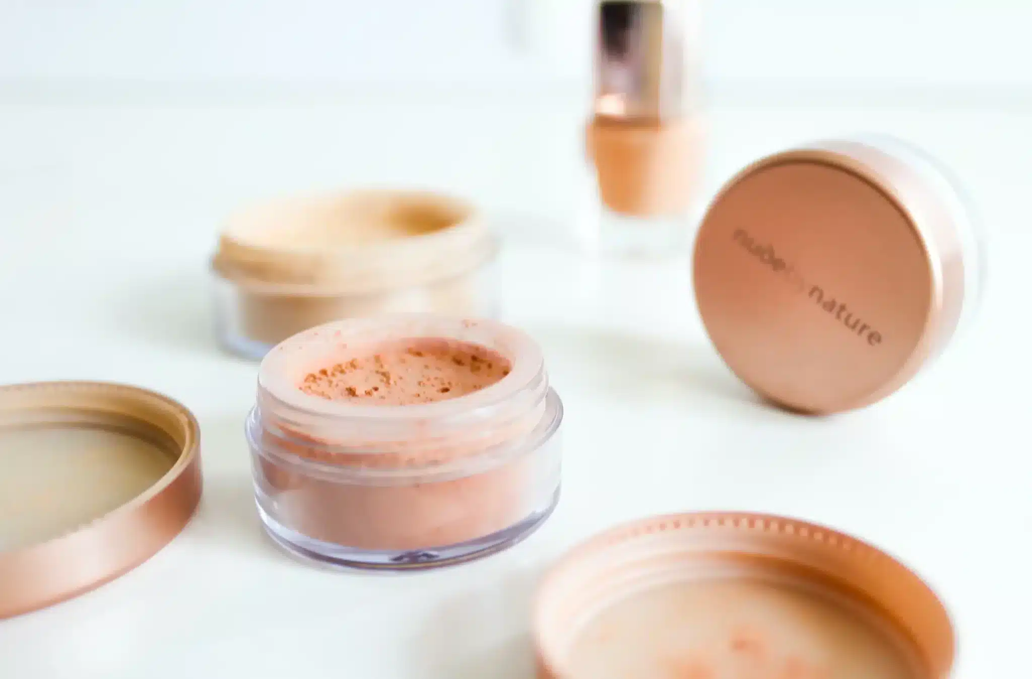 A soft-focused image showcasing an elegant display of cosmetic products with a clear emphasis on natural beauty. In the foreground, an open jar of loose powder in a delicate pink shade sits beside its matching lid, with the brand 'nude by nature' etched on it. The background features a blurred foundation bottle, all set against a crisp white backdrop.