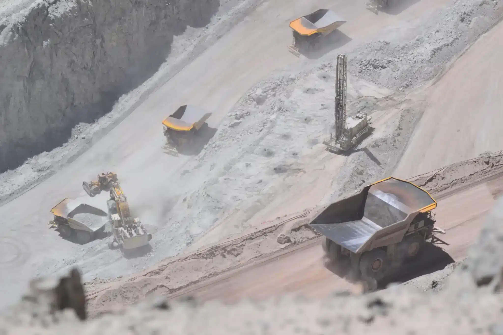 An active open-pit mining operation with large machinery at work. Several heavy dump trucks with yellow beds are visible transporting materials, while an excavator loads one of them. A drilling rig stands in the background amidst a landscape of exposed earth and rock.