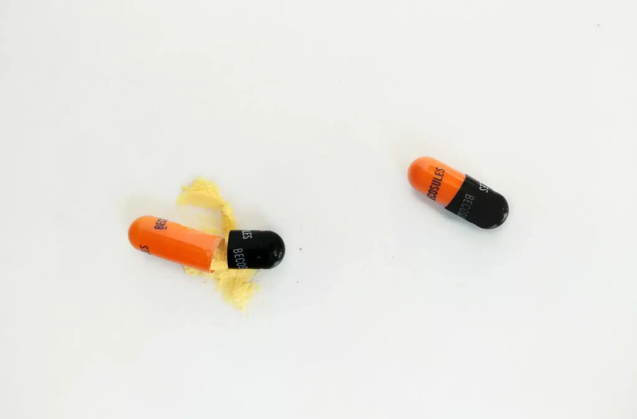 An image of two capsules on a white background, one capsule is partially opened with its orange and black contents spilled out, resembling powder. The other intact capsule has a similar color pattern. Both are marked with 'B6 300mg' signifying their dosage and potential vitamin content.