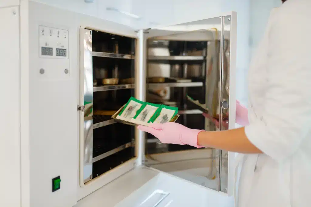 A person in a lab coat with pink gloves placing a tray with green-packaged products into a tray dryer with a digital control panel.