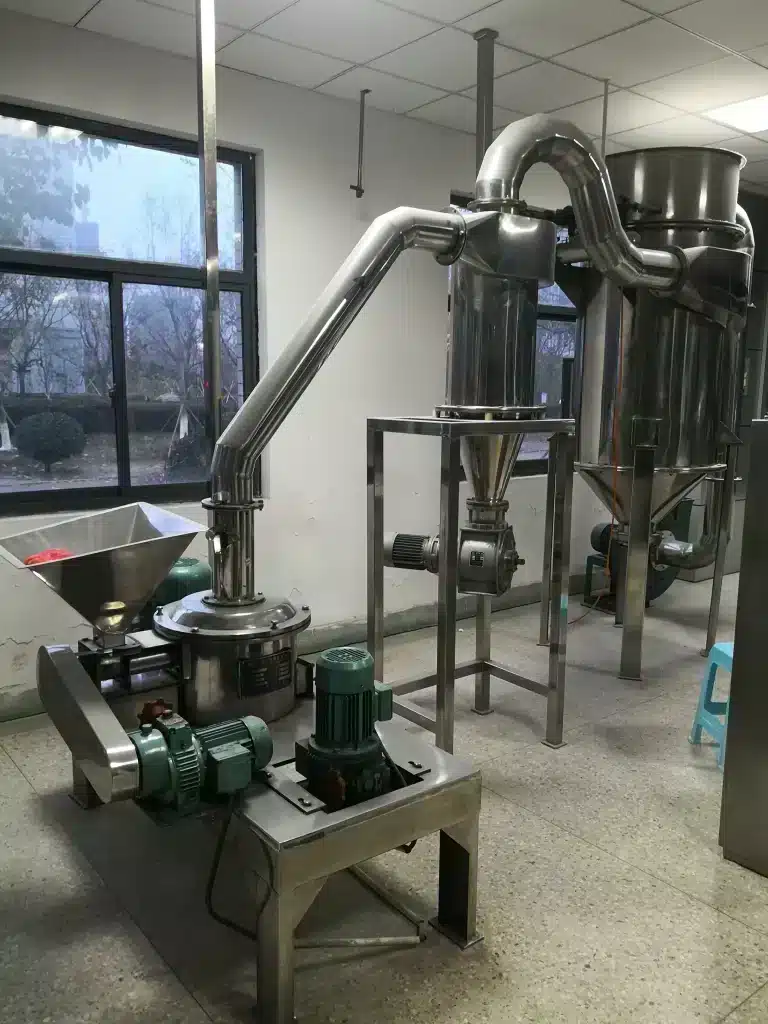 Industrial room with an impact pulverizer, showing a compact machine with a feeding hopper and two motors mounted on a metal frame. Pipes extend from the top, connecting to a larger vertical cylindrical chamber, likely for pulverizing and processing materials.
