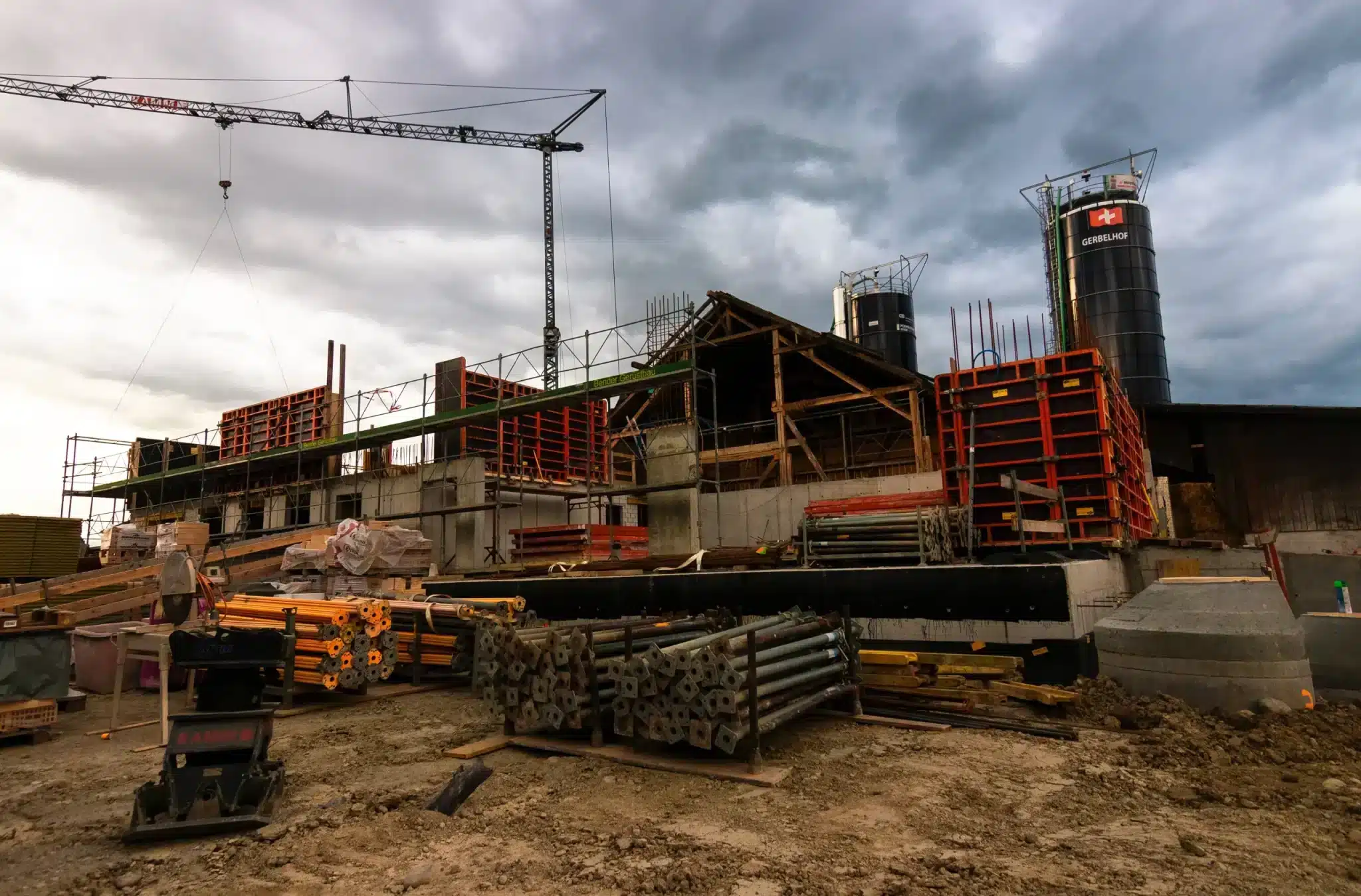 A construction site under a moody sky, with a large crane towering above and various building materials strewn about. The skeleton of a building with orange and red formwork is taking shape, with concrete silos emblazoned with the Swiss flag in the background.