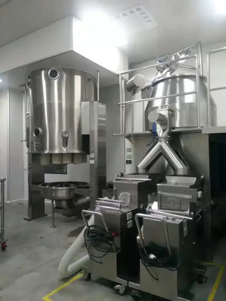 A sophisticated vertical fluidized bed dryer dominates an industrial room with shiny stainless steel surfaces. To the left, a tall cylindrical structure with various ports and gauges is present, indicative of the fluidizing chamber. In the foreground, complex machinery with tubing and control panels is seen, likely for material handling and processing within the dryer system.