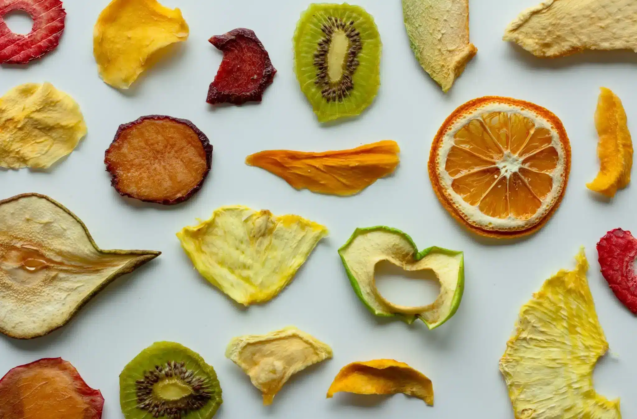 An assortment of vibrant dried fruit slices spread out against a white background. The collection includes strawberries, apples, oranges, kiwis, and pears among others, showcasing a variety of shapes and rich colors, with one piece cut into a heart shape.