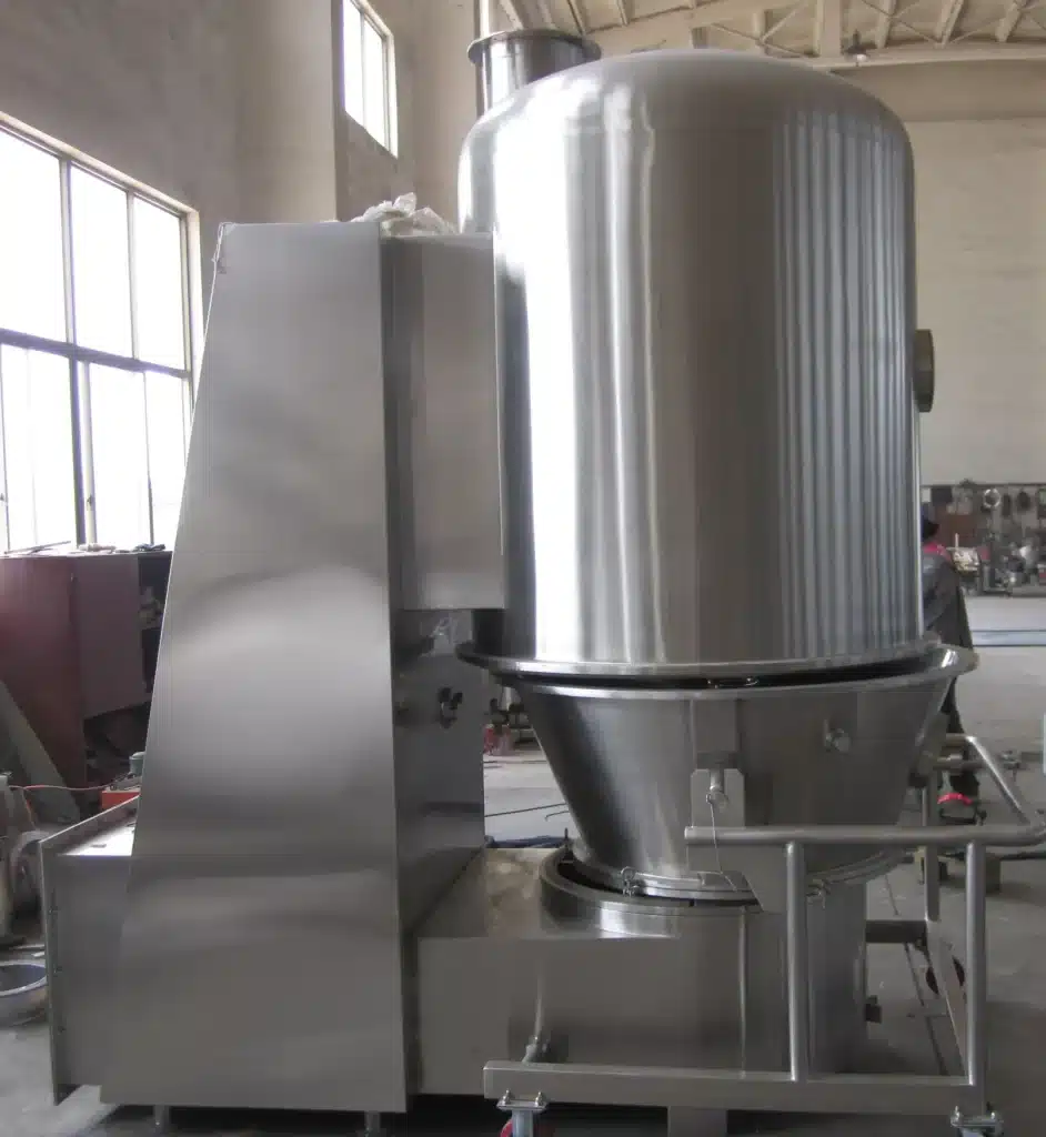 A large Vertical Blending Fluidized Bed Dryer stands in an industrial workshop, featuring a tall stainless steel vessel with a conical base and central pipe, on a metal frame with wheels, suggesting mobility and flexibility within the manufacturing space.