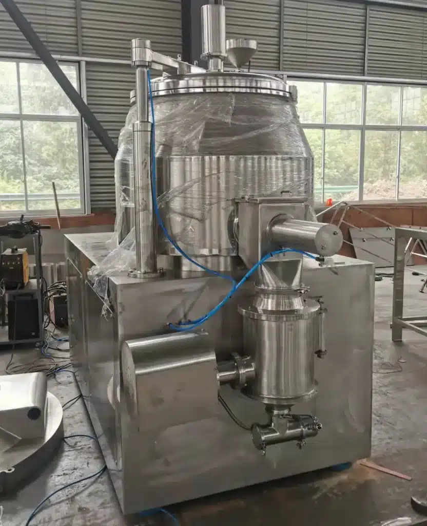 A Wet Rapid Mixing Granulator featuring a large mixing bowl with a clear lid, stainless steel construction, and various inlet pipes, set in an industrial manufacturing space with natural light.