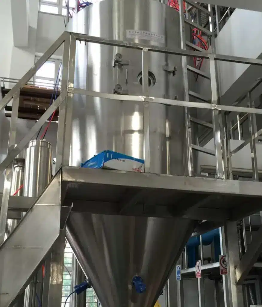 A Closed Circulation Spray Dryer set up in an industrial environment, featuring a tall cylindrical body with inspection windows and a conical bottom. The structure is supported by a metal frame with stair access, highlighting its closed-loop design for safe, controlled drying processes.