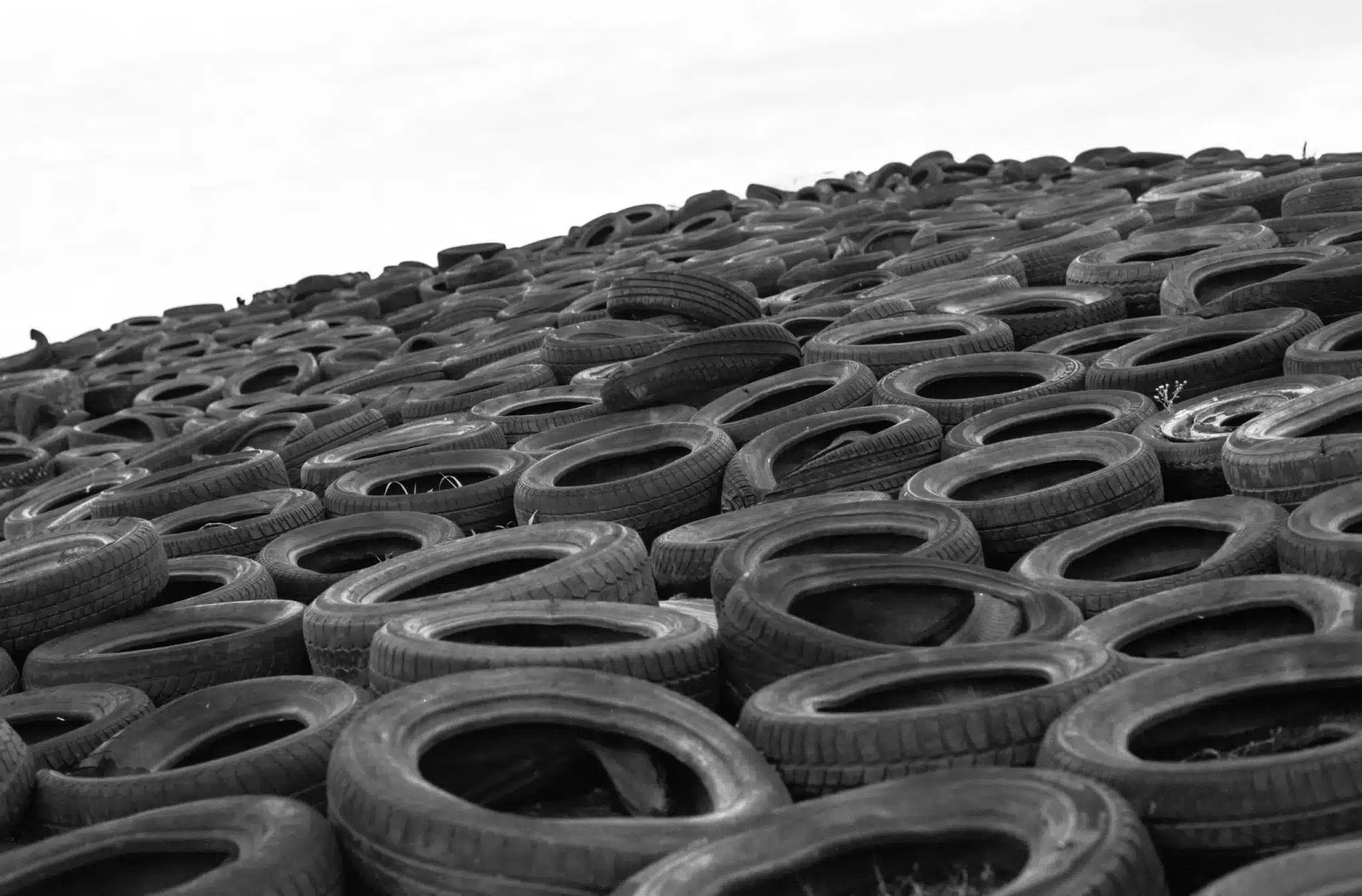 A black and white photo capturing a vast pile of discarded tires extending into the distance, symbolizing issues of waste and recycling in modern society. The image shows the varied sizes and worn textures of the tires, starkly piled against a pale sky.