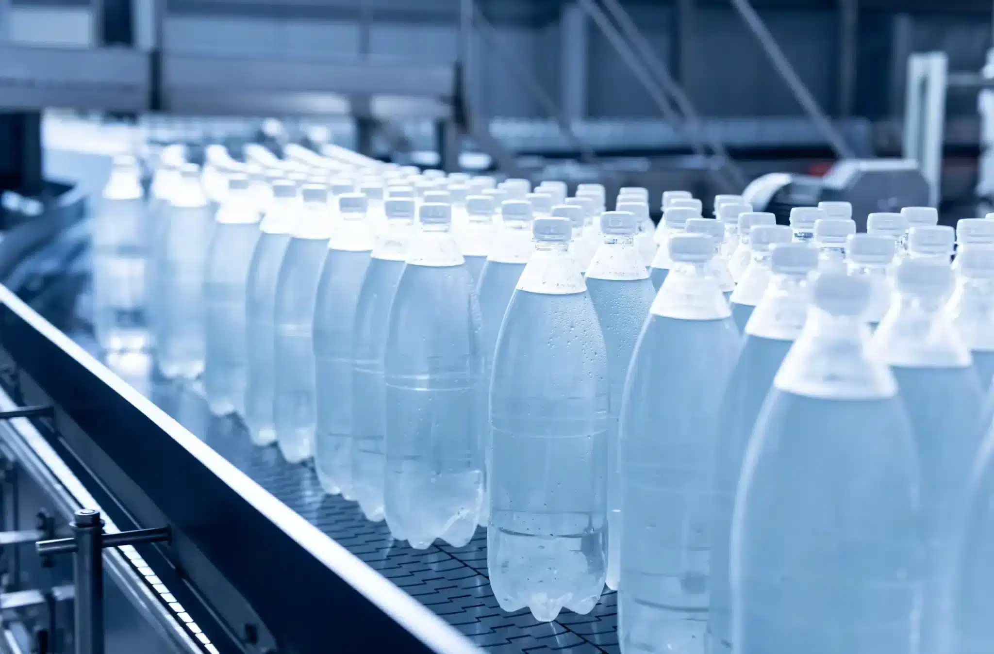A production line in a factory with a row of clear, empty plastic bottles moving along a conveyor belt. The image captures the industrial process of packaging with a cool, blue tone, emphasizing the bottles' uniformity and the automated manufacturing environment.