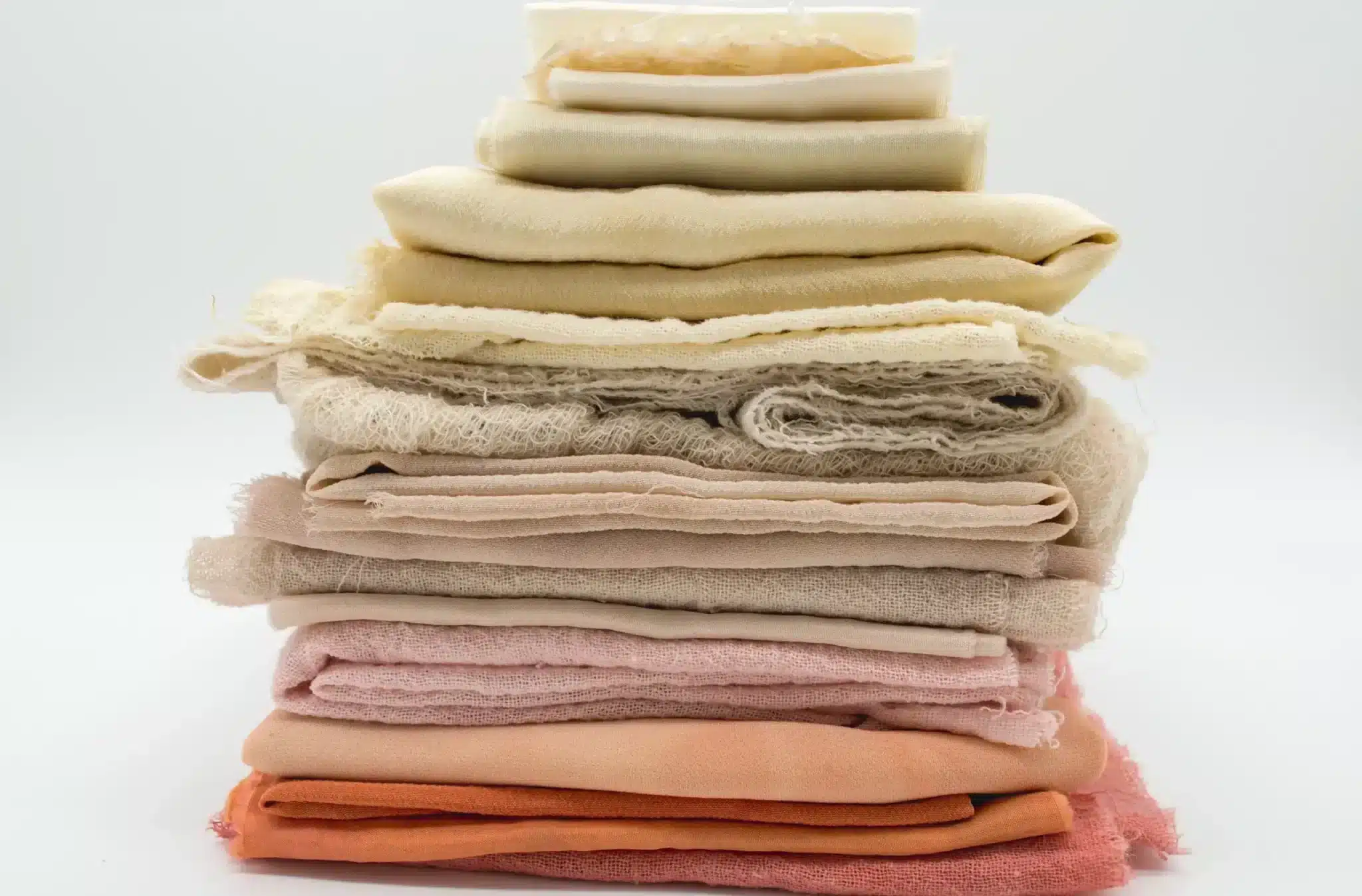 A neatly stacked pile of fabrics in a gradient of colors, from cream and beige to dusty pink and terracotta, creating a soothing palette. The textures range from smooth and fine to more rustic and loosely woven, suggesting a variety of uses in textile design.