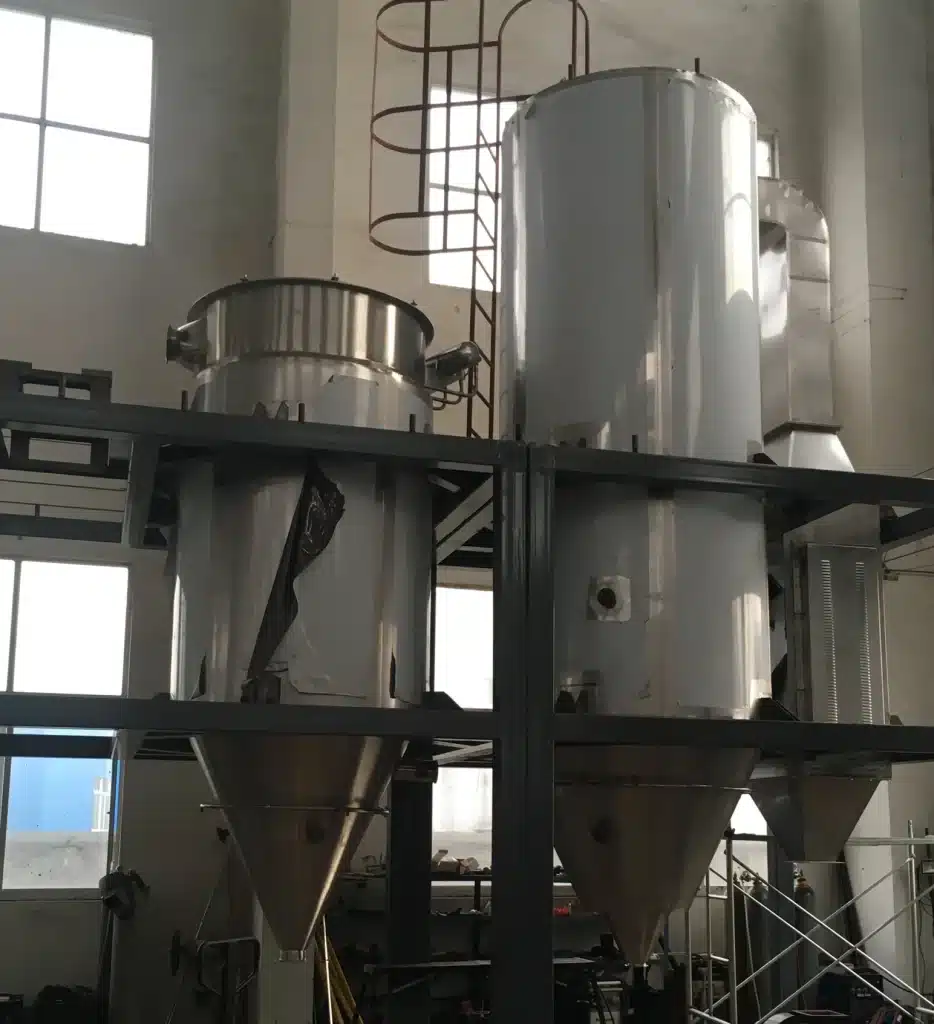 Industrial Pressure Spray Dryer installation in a warehouse setting, with a tall, cylindrical steel body and conical base. The machinery includes a platform with railings and is surrounded by various tools and components, illustrating a setup or maintenance phase.