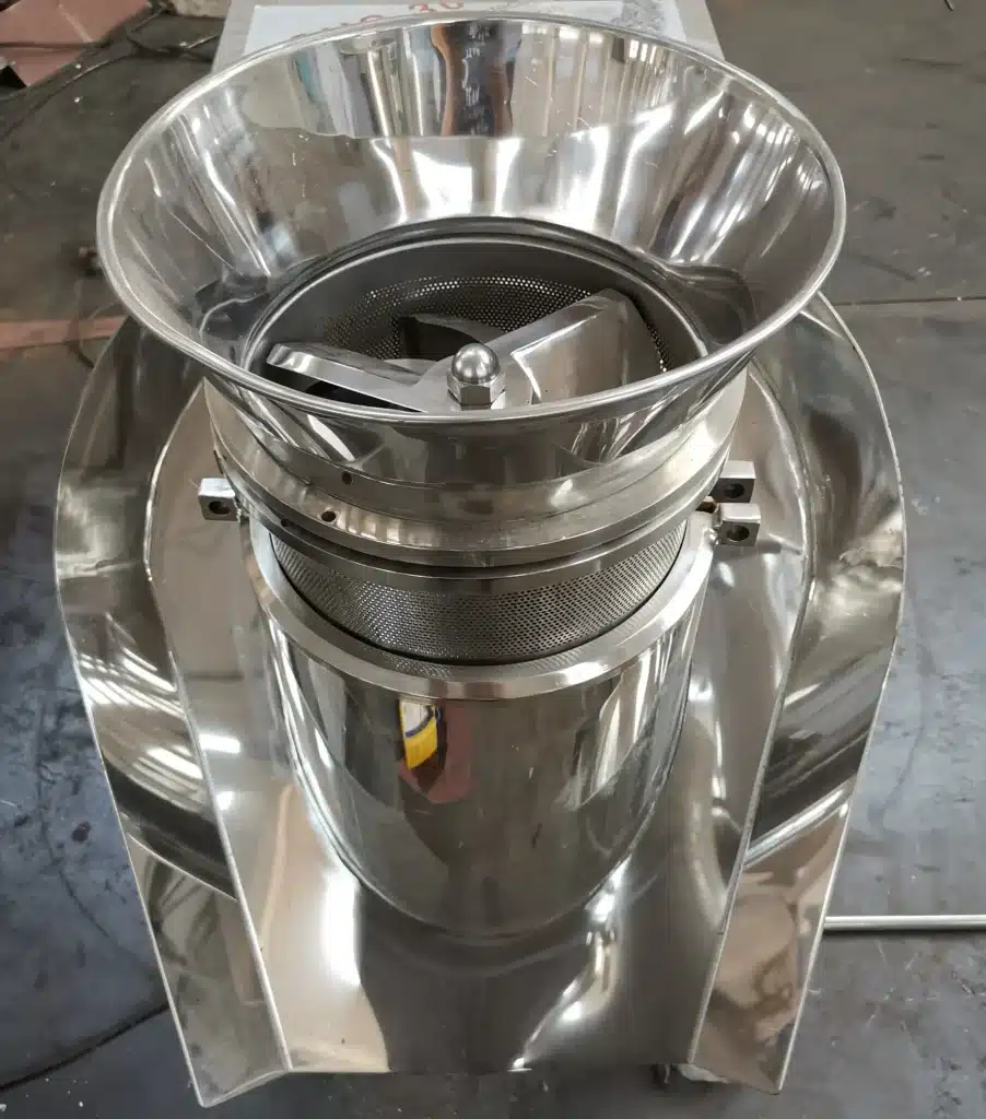 A stainless steel Basket Granulator featuring a mesh drum within a funnel-shaped holder, situated in an industrial workspace.