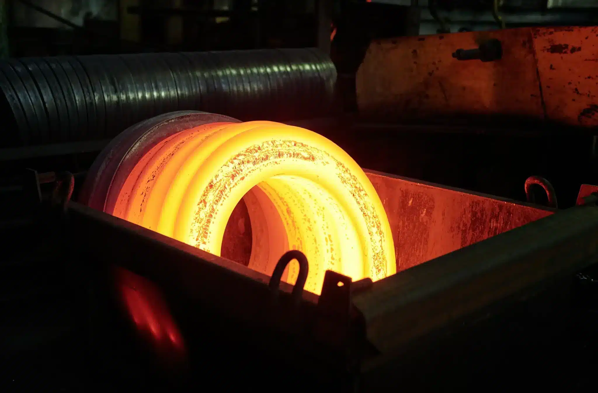 A hot steel coil glowing in an intense orange hue as it's being processed in an industrial setting. The heat emanating from the metal is palpable, highlighting the raw power of manufacturing and metallurgy.