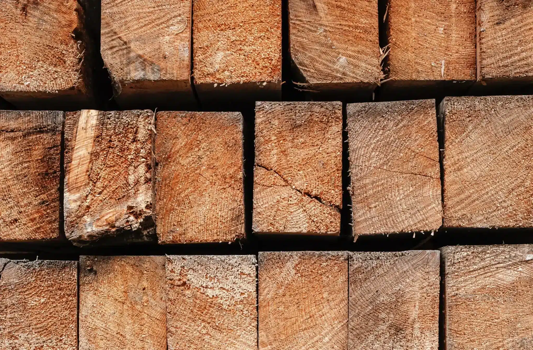 The end-grain of a stack of freshly cut lumber, showing the natural patterns and rings of the wood. The warm brown tones and textures are highlighted by sunlight, indicating the material's readiness for use in construction or carpentry.
